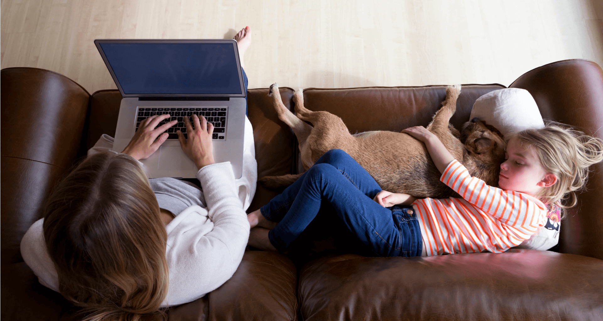 mother using apple laptop on couch while daughter cuddles with dog next to her
